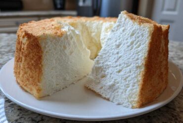 A tall, fluffy slice of angel food cake with a golden crust sitting next to the rest of the cake on a white plate.