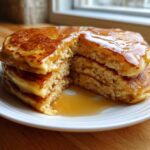 A stack of three fluffy discard pancakes, cut open to show the texture, generously covered in maple syrup.