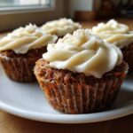 Four delicious carrot cake muffins topped with thick cream cheese frosting, sitting on a white plate.