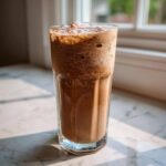A tall glass filled with thick, blended frozen hot chocolate sitting on a marble surface near a window.