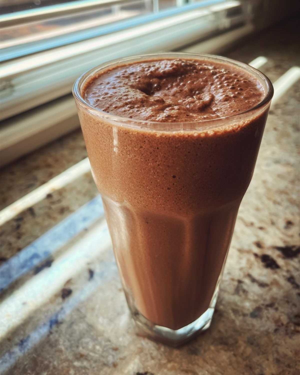 A tall glass filled with thick, blended frozen hot chocolate sitting on a granite countertop near a window.