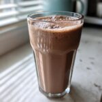 A tall glass filled with frothy, blended frozen hot chocolate sitting on a white counter near a window.