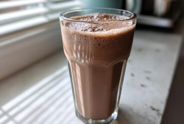A tall glass filled with frothy, blended frozen hot chocolate sitting on a white counter near a window.
