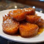 A close-up of several thick, round slices of glistening candied yams piled on a white plate, coated in a rich, dark syrup.