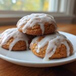 A stack of three soft, spiced persimmon cookies topped with a thick white glaze, resting on a white plate.