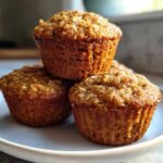 Three golden brown banana oatmeal muffins stacked on a light gray plate, showing visible oats on top.