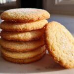 A stack of five golden brown sugar cookie rounds, topped with sparkling sugar, with one leaning against the pile.