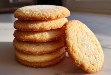 A stack of five golden brown sugar cookie rounds, topped with sparkling sugar, with one leaning against the pile.