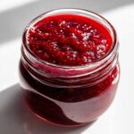 Close-up of a glass jar filled with rich, dark red homemade raspberry jam, showing texture and seeds.