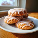 A stack of soft, orange persimmon cookies drizzled with white icing, sitting on a white plate near a window.