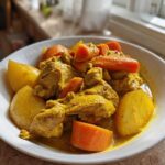 A white bowl filled with vibrant yellow Jamaican curry chicken, potatoes, and carrots, ready to eat.