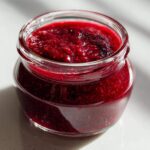 Close-up of a small glass jar filled with thick, vibrant red raspberry jam, sitting on a bright surface.