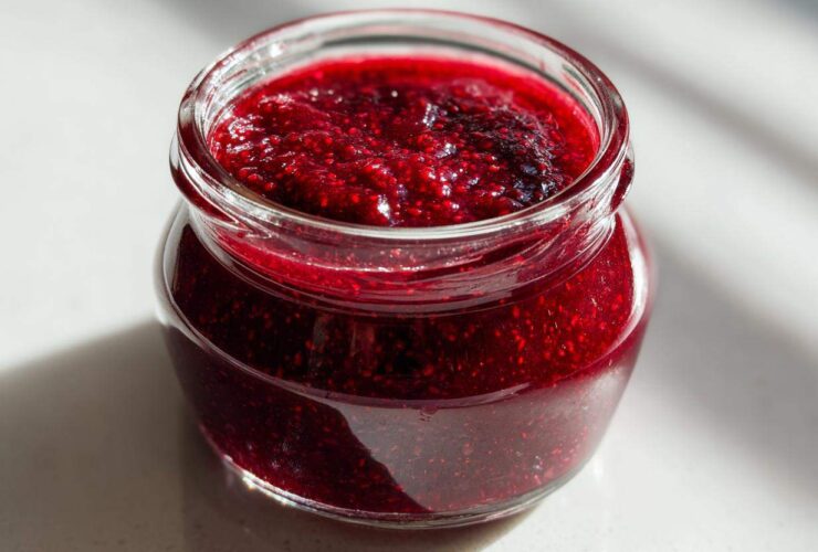 Close-up of a small glass jar filled with thick, vibrant red raspberry jam, sitting on a bright surface.