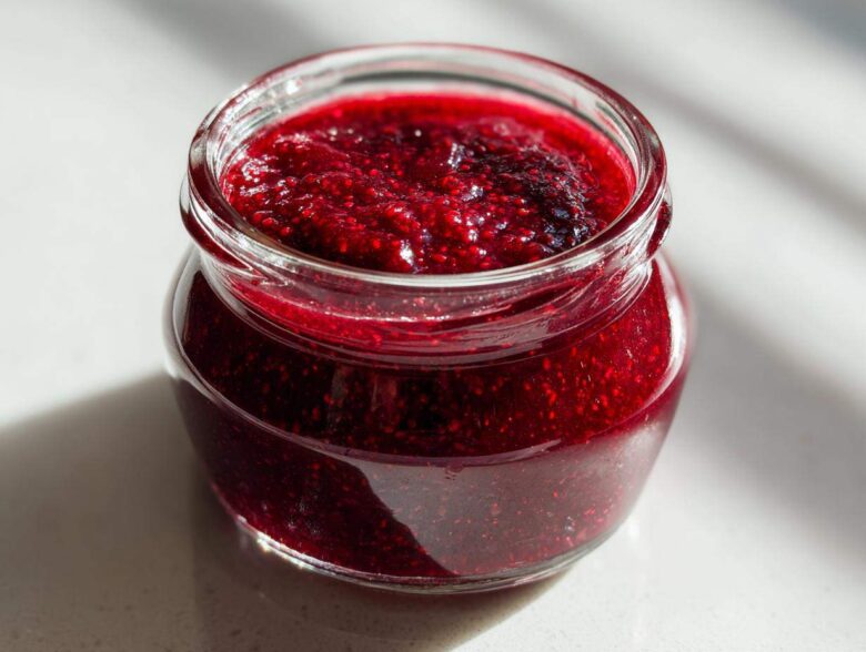 Close-up of a small glass jar filled with thick, vibrant red raspberry jam, sitting on a bright surface.