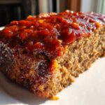 A thick slice of moist meatloaf topped with a shiny, reddish-brown glaze, sitting on a white plate.