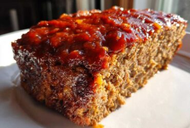 A thick slice of moist meatloaf topped with a shiny, reddish-brown glaze, sitting on a white plate.