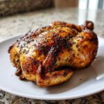 A whole, perfectly seasoned and juicy roasted chicken with crispy, dark brown skin resting on a white plate.