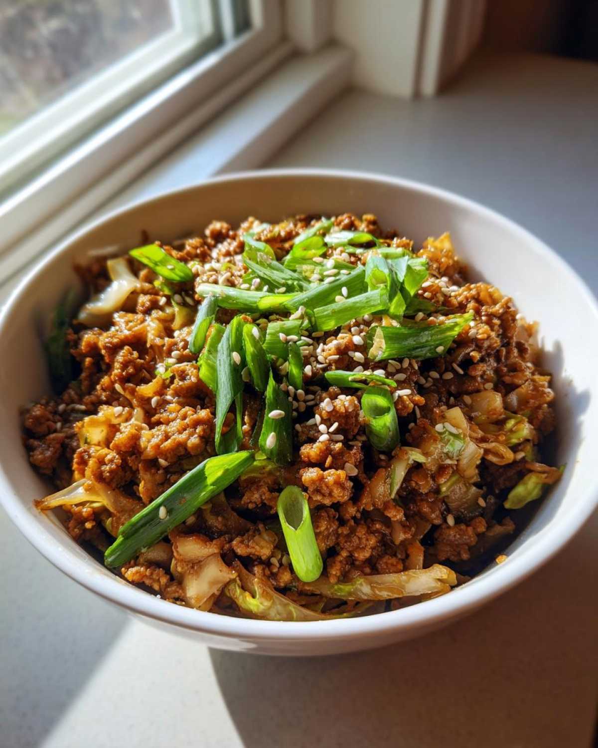 A close-up of a white bowl filled with keto egg roll in a bowl, topped with green onions and sesame seeds.