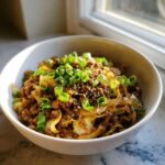 A serving of keto egg roll in a bowl with ground meat and cabbage, topped generously with sliced green onions and sesame seeds.