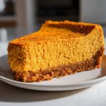 A rich, vibrant orange slice of keto pumpkin cheesecake with a thick graham cracker-style crust, served on a white plate.