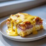 A decadent slice of eggs benedict casserole layered with bread, ham, egg, and covered in rich hollandaise sauce.