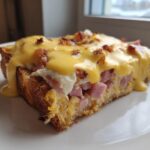A single serving slice of make-ahead eggs benedict casserole topped generously with bright yellow hollandaise sauce and bacon bits.