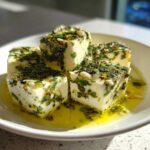 Four cubes of creamy marinated cheese generously coated in green herbs and submerged in golden olive oil.