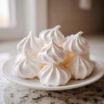 A close-up of several perfectly piped, bright white meringue cookies stacked on a small white plate.