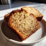 Close-up of three moist slices of banana bread with dark specks on a white plate, highlighted by bright sunlight.