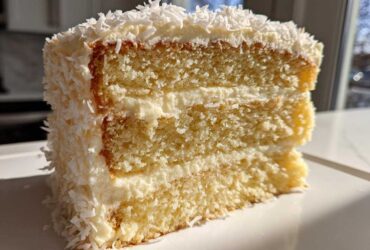 Close-up of a moist slice of layered coconut cake recipe covered in white frosting and shredded coconut flakes.
