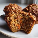 Close-up of a moist morning glory muffin with a bite taken out, revealing shredded carrots and nuts inside.