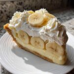 A decadent slice of no bake banana cream pie featuring a graham cracker crust, thick custard, and topped with whipped cream and fresh banana slices.