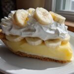 A generous slice of no bake banana cream pie featuring a graham cracker crust, thick vanilla filling, and topped with whipped cream and fresh banana slices.