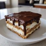 A square slice of no-bake chocolate eclair cake featuring layers of graham crackers, cream filling, and a thick chocolate glaze dripping down the sides.