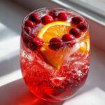 Close-up of a glass filled with bubbling red non-alcoholic christmas punch, garnished with fresh cranberries and an orange slice.