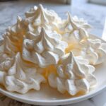 A close-up of several light, white, piped meringue cookies stacked on a white plate.