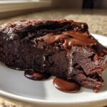 A dense slice of flourless chocolate cake topped with dripping chocolate ganache, served on a white plate.