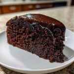 A close-up of a dense slice of flourless chocolate cake topped with glossy, dripping chocolate ganache.
