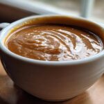Close-up of thick, rich turkey gravy with swirls in a white ceramic mug, set near a window.