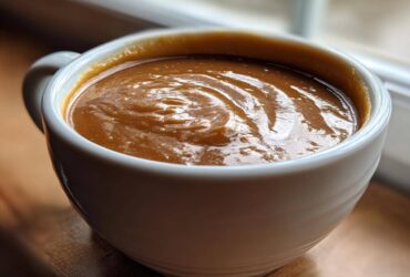 Close-up of thick, rich turkey gravy with swirls in a white ceramic mug, set near a window.