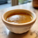 Close-up of rich, brown turkey gravy served in a small white fluted ramekin on a marble surface.