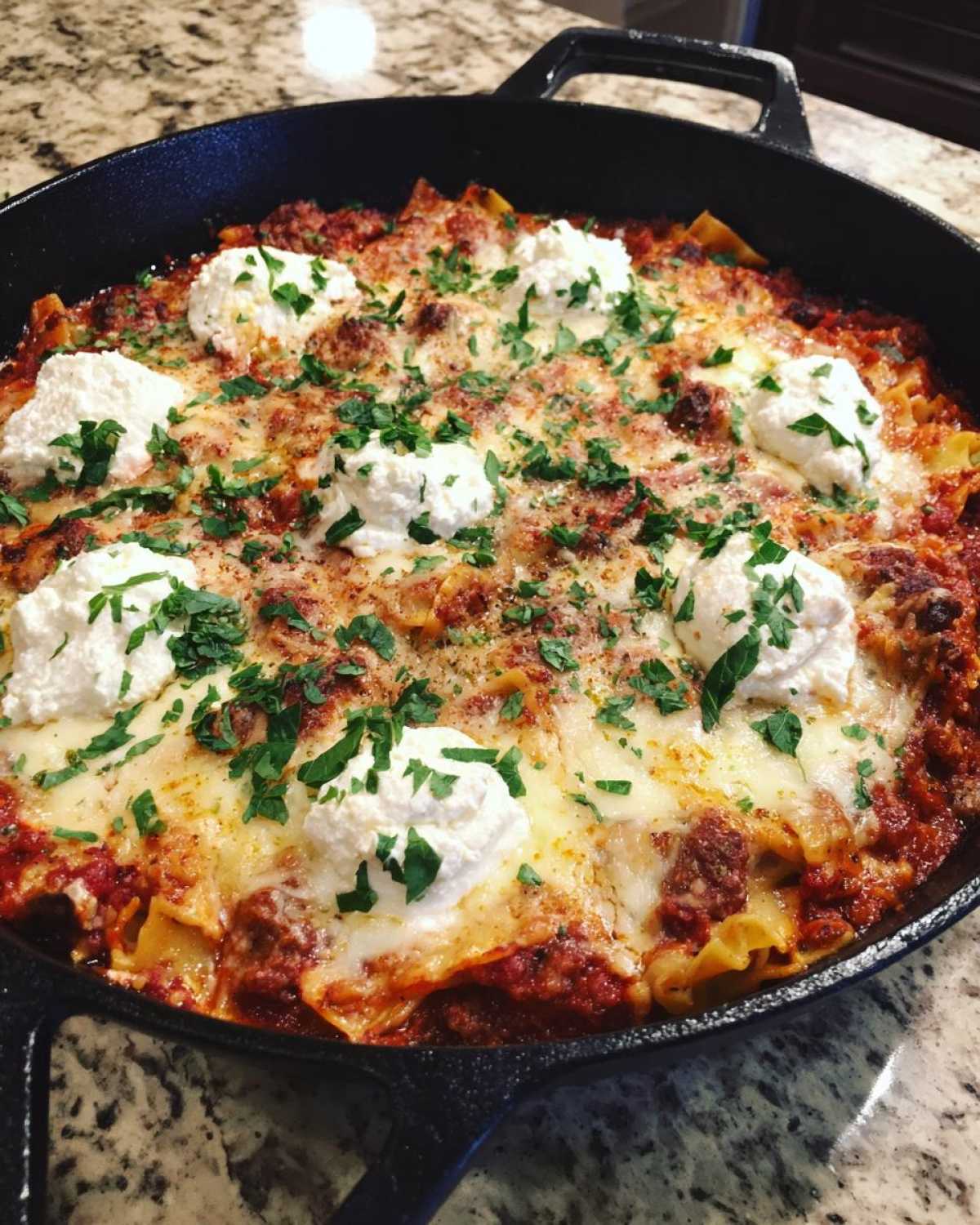 Close-up of a bubbling skillet lasagna in a cast iron pan, topped with melted cheese, dollops of ricotta, and fresh parsley.