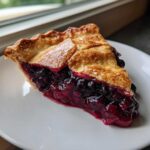 A close-up of a thick slice of homemade blueberry pie recipe with a flaky golden crust and rich, juicy filling.