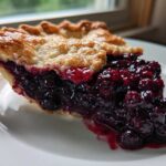 Close-up of a thick slice of blueberry pie recipe with a flaky crust and rich, juicy blueberry filling.