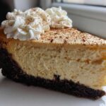 A close-up of a rich slice of eggnog cheesecake recipe with a dark crust, topped with whipped cream and cinnamon.