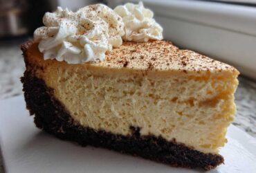 A close-up of a rich slice of eggnog cheesecake recipe with a dark crust, topped with whipped cream and cinnamon.