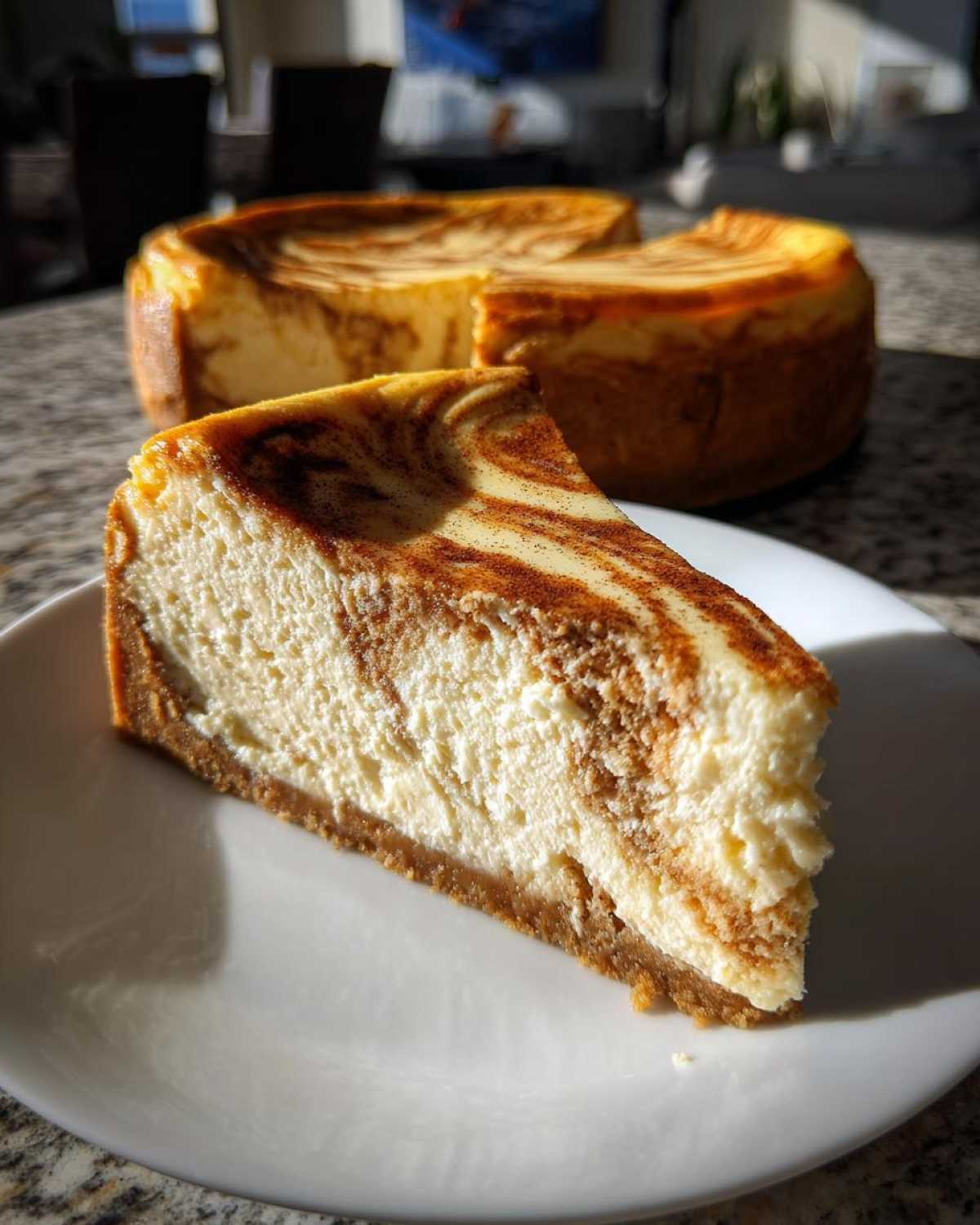 A perfect slice of snickerdoodle cheesecake with a cinnamon swirl topping sits on a white plate, with the rest of the cake blurred in the background.