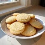 A pile of freshly baked, soft and chewy sugar cookies stacked on a white plate near a sunny window.