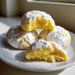 Four bright yellow lemon crinkle cookies heavily dusted with powdered sugar, one cookie is broken open to show the soft interior.