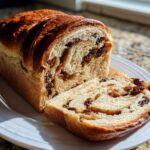 A freshly baked loaf of soft raisin bread, partially sliced, displaying swirls of cinnamon and plump raisins.
